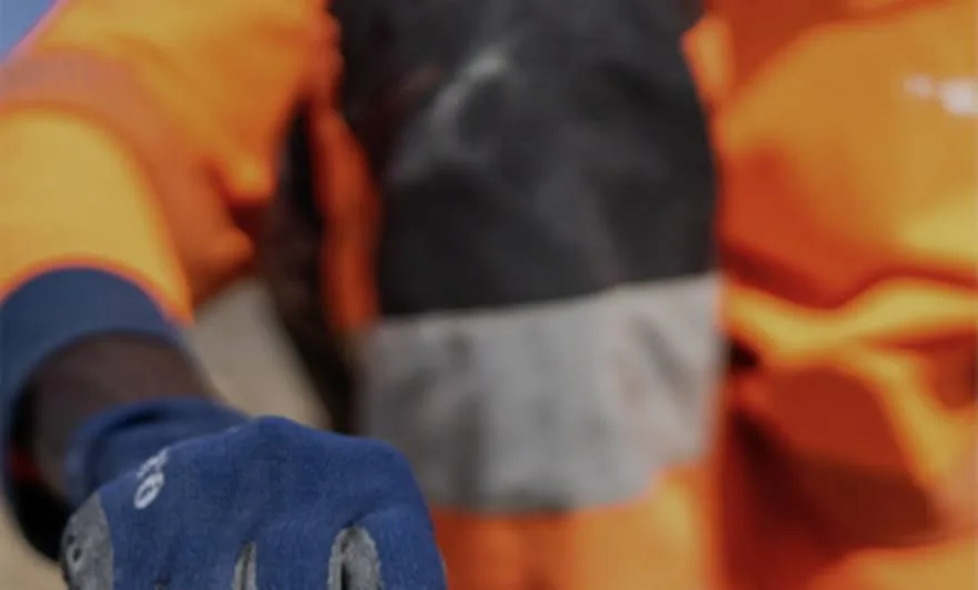 Gros plan sur le buste d'un opérateur en tenue haute visibilité orange à bandes réfléchissantes, tendant une main gantée de bleu vers l'objectif.