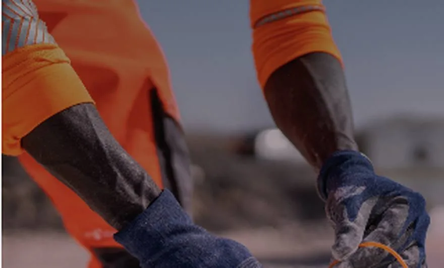 Gros plan sur les avant-bras et les mains gantées de deux opérateurs en tenue haute visibilité orange, se passant un objet sur une aire de chantier.