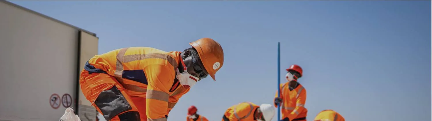 A blasting operative in high-visibility orange clothing, helmet and protective mask leans over a charge on the ground; other team members work in the background under a clear sky.