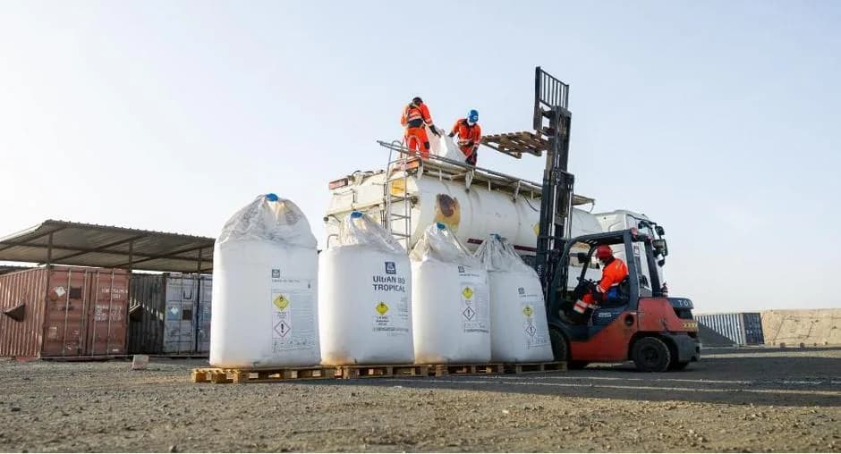 Storage area under a shed: large white bulk bags of product lined up on pallets, a red forklift and a technical building.