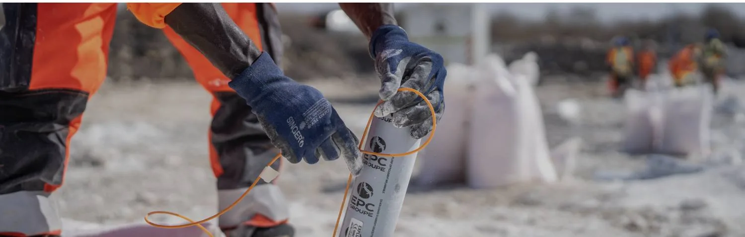 The gloved hands of an operator place a cartridge of explosive into a blasthole, on light-coloured rock.